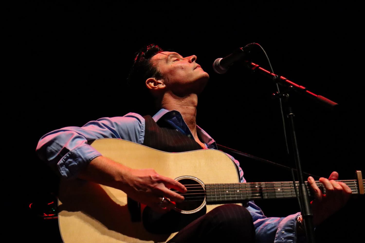 Man playing acoustic guitar and singing into microphone on a dark stage with red lighting.