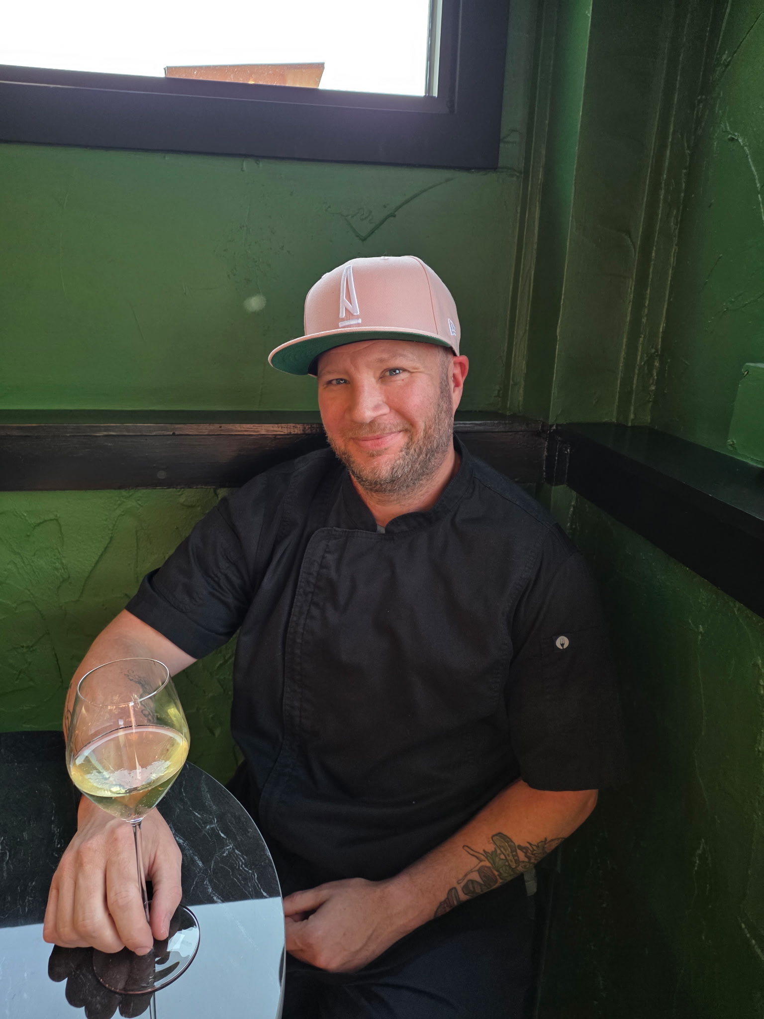 Man in a black chef jacket and pink cap sitting at a black table holding a glass of white wine.
