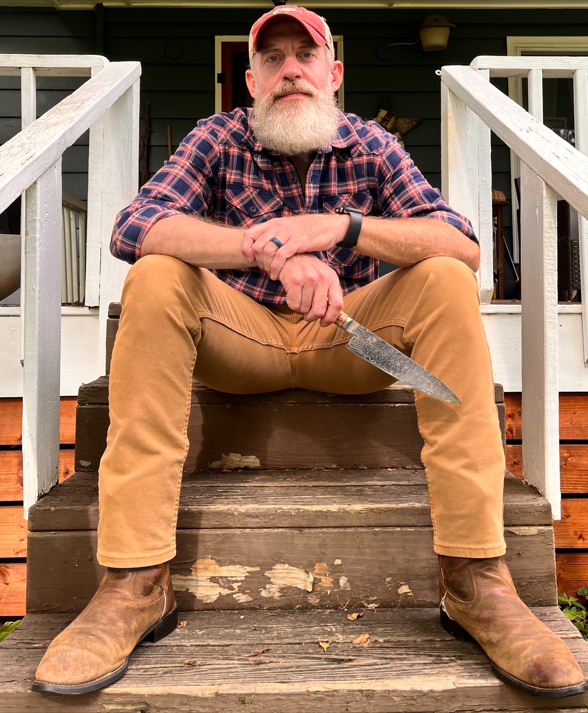 Jon Sawrie Headshot - Supper Club Bearded man in red cap, plaid shirt, tan pants, and brown boots sitting on wooden steps holding patterned knife.