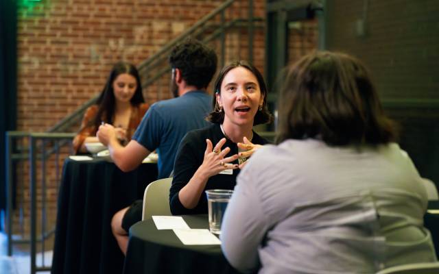 Two pairs of people seated at black-covered tables in a brick-walled room, engaged in conversation.