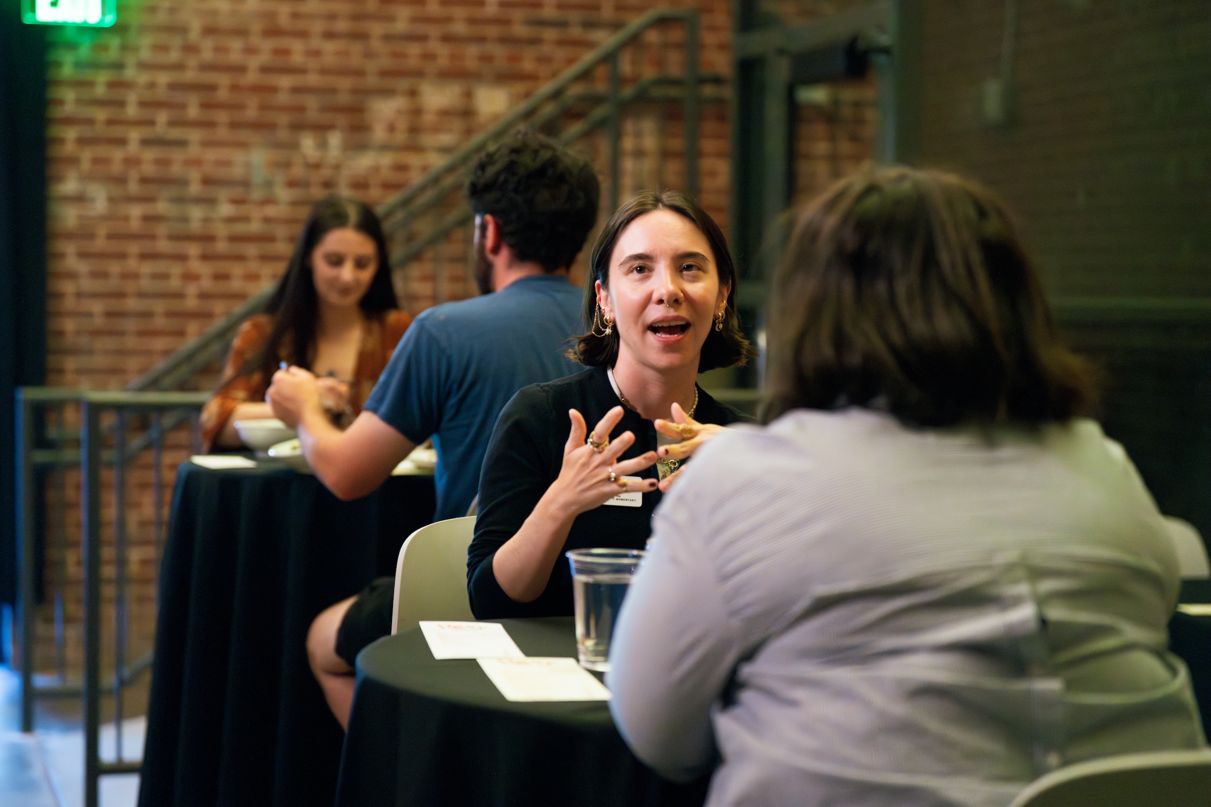 Two pairs of people seated at black-covered tables in a brick-walled room, engaged in conversation.