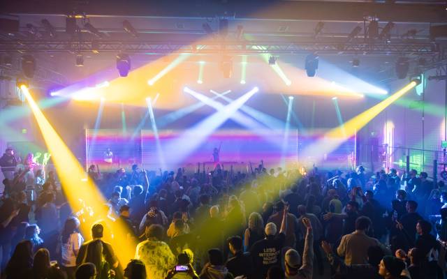 Indoor concert with DJ and vibrant colorful stage lighting illuminating the crowd.