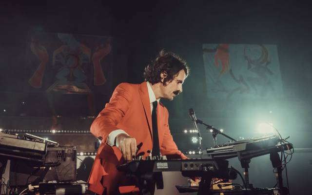 Man in orange suit performing electronic music on stage with keyboard and mixer.