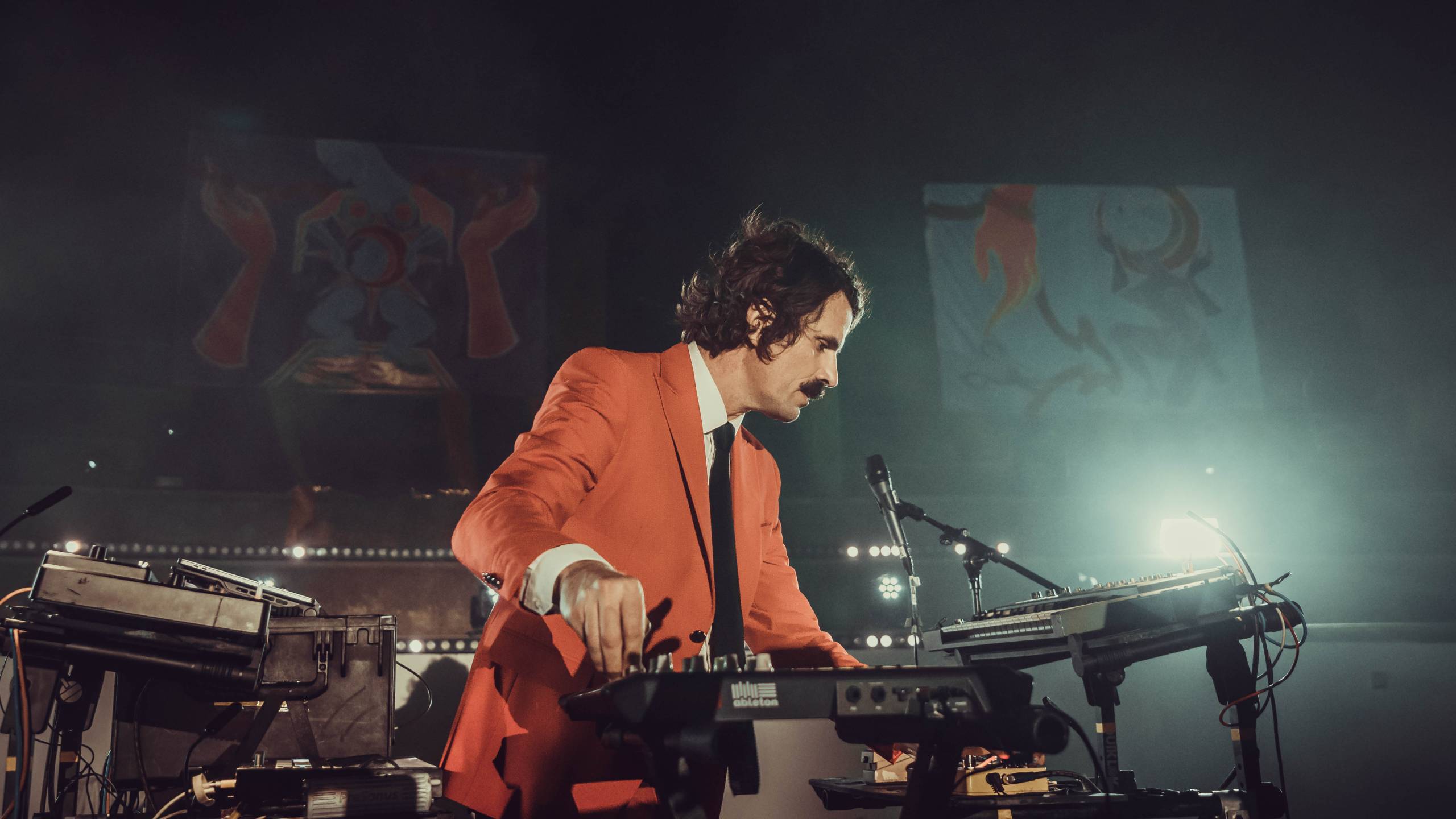 Man in orange suit performing electronic music on stage with keyboard and mixer.