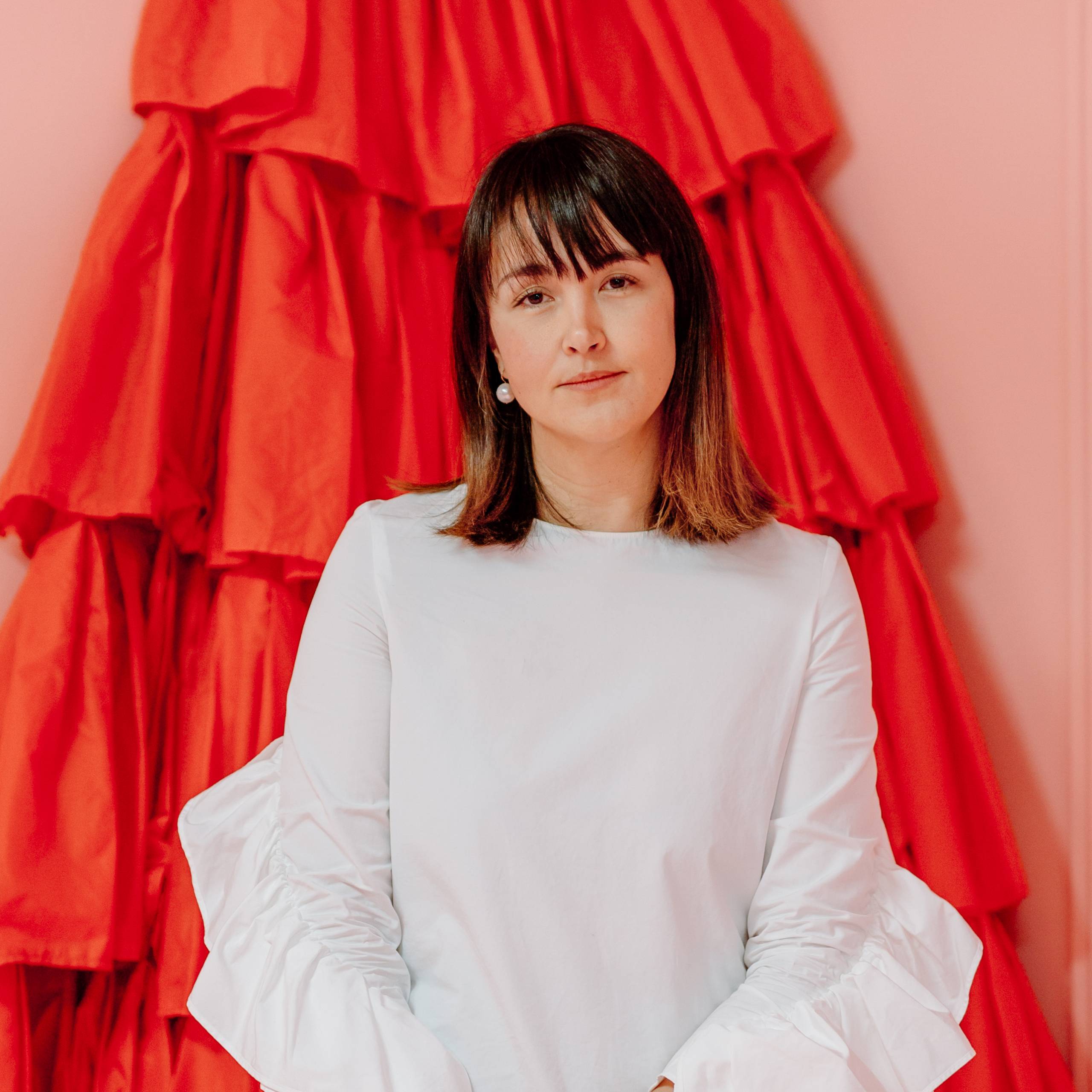 Danielle Hatch Headshot (1) (1) Woman with shoulder-length brown hair and bangs wearing a white blouse in front of red ruffled fabric.