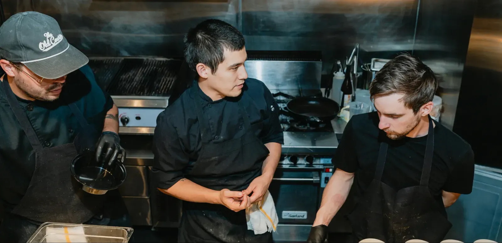 Three chefs in black uniforms prepare food in a professional kitchen with cooking equipment.