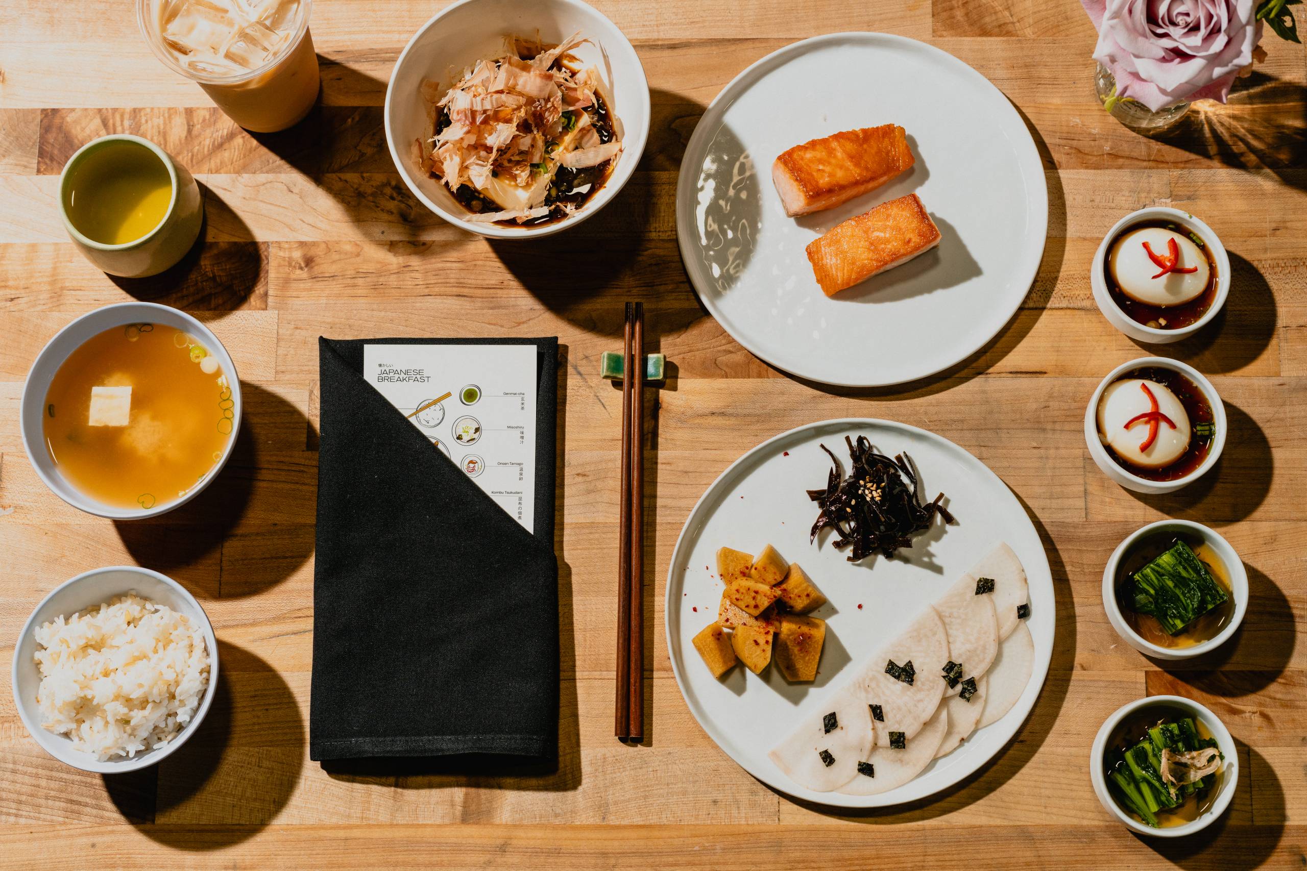 Japanese breakfast with grilled salmon, rice, miso soup, tofu, soft boiled eggs, and vegetables on a wooden table.