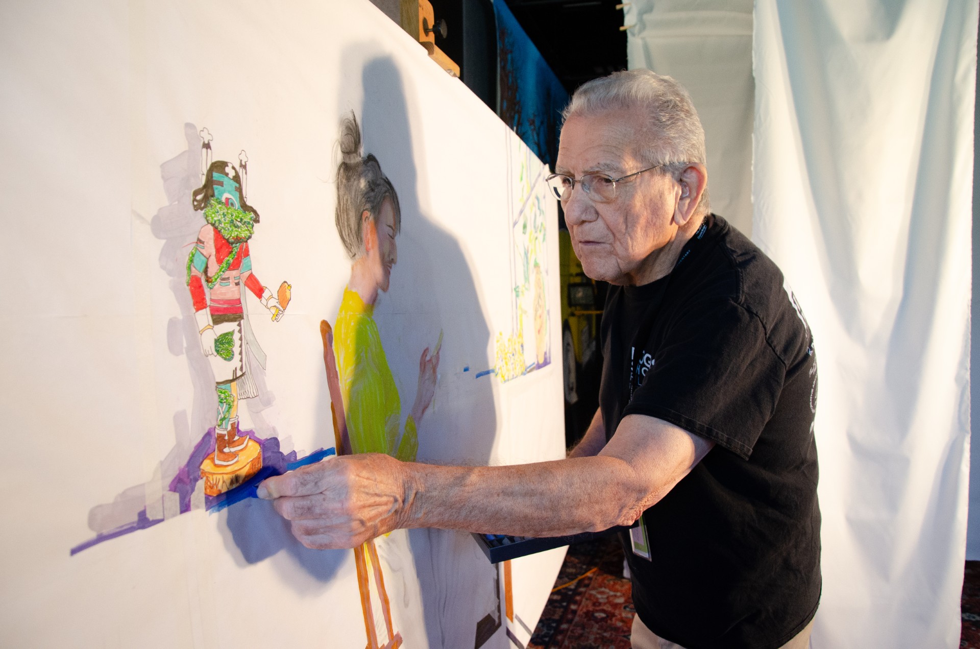 Elderly man wearing glasses painting a detailed mural featuring a woman and decorative figure.