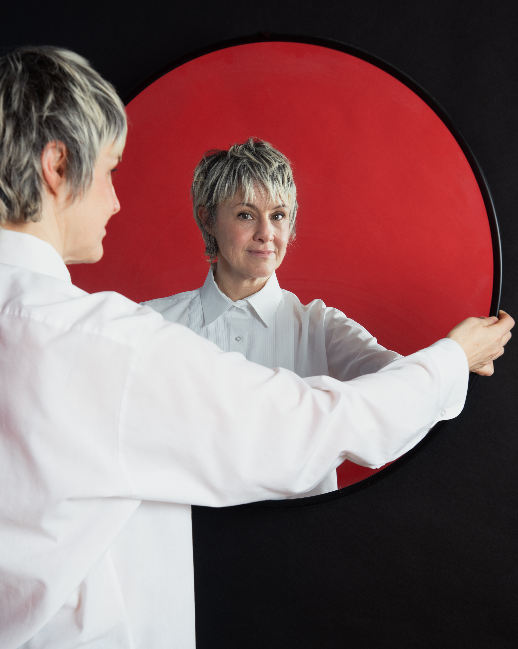 Person with short light hair holding a round mirror reflecting their face and white shirt against red and black backgrounds