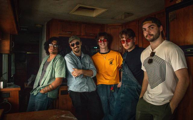 Five casually dressed young men standing together in a room with wooden cabinets.