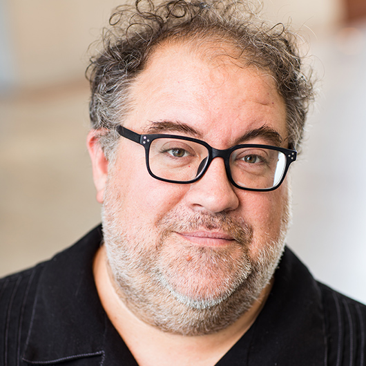 Stephen-Moore-Photo Middle-aged man with curly hair, beard, and black rectangular glasses wearing a dark shirt.