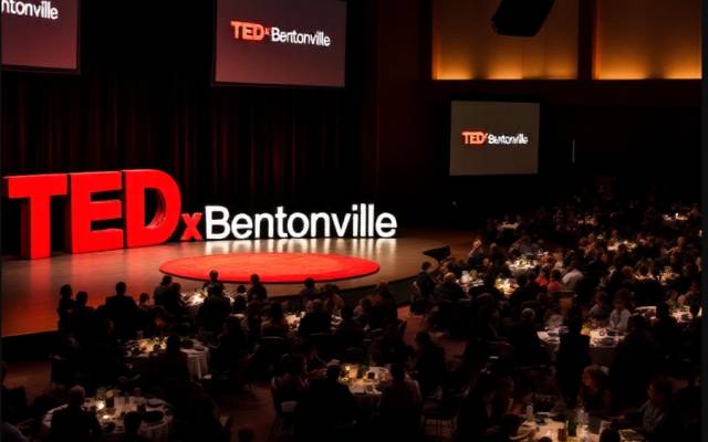 TEDxBentonville event with audience seated at round tables facing stage and screens.