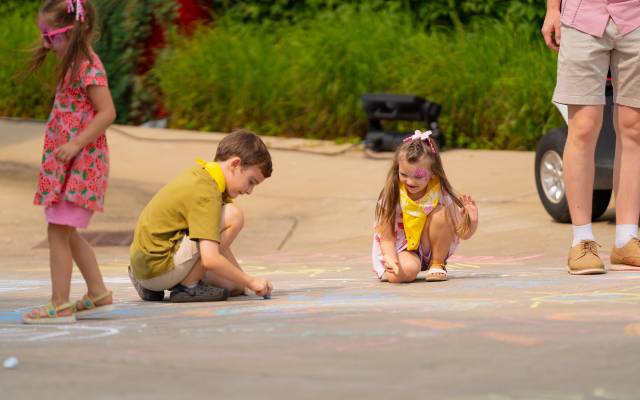 Children drawing with colorful chalk on pavement outdoors near an adult's legs.