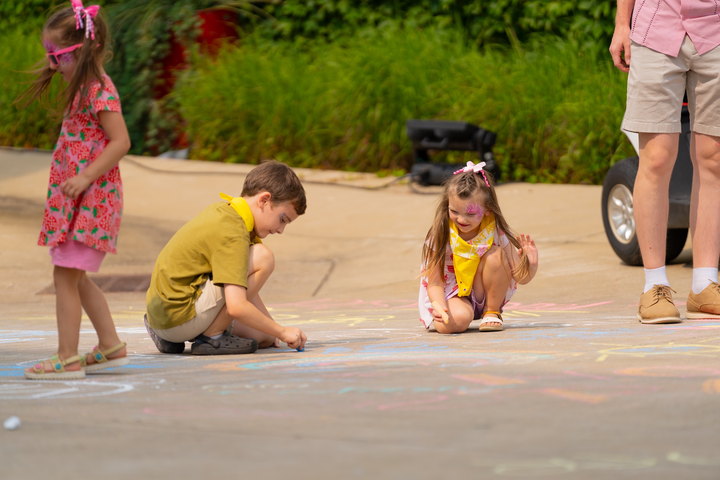 Children drawing with colorful chalk on pavement outdoors near an adult's legs.