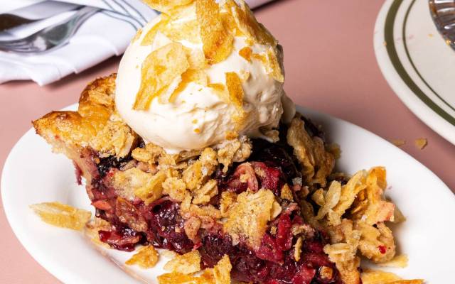 Slice of fruit pie topped with vanilla ice cream and crispy flakes on a white plate.