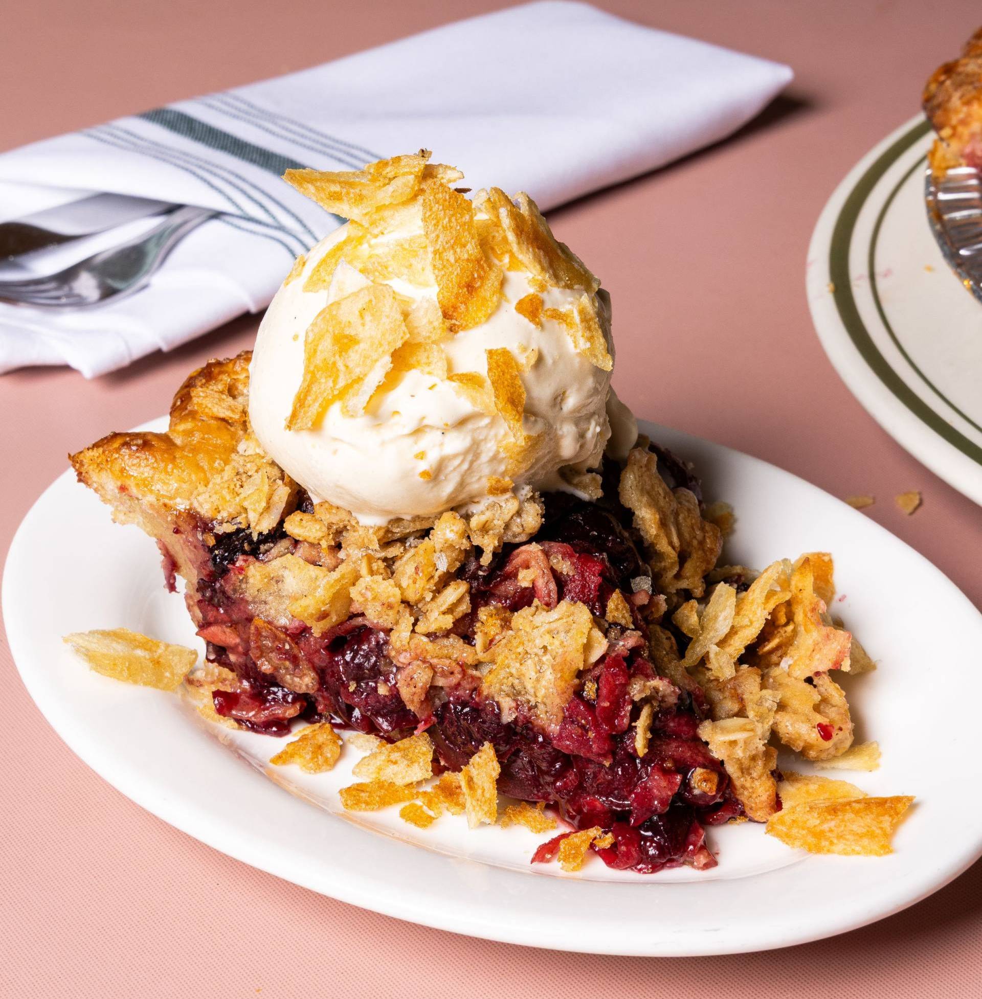 Slice of fruit pie topped with vanilla ice cream and crispy flakes on a white plate.