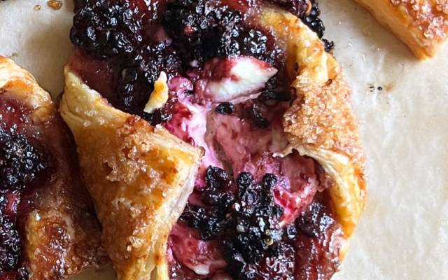 Baked pastries with golden crust filled with cream and dark berry topping on parchment paper