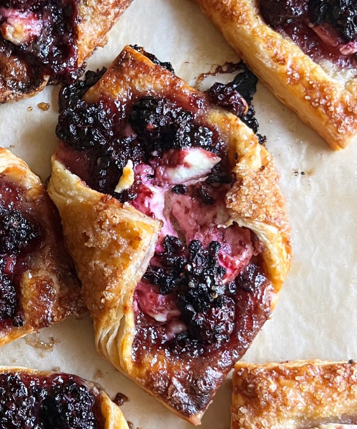 Baked pastries with golden crust filled with cream and dark berry topping on parchment paper