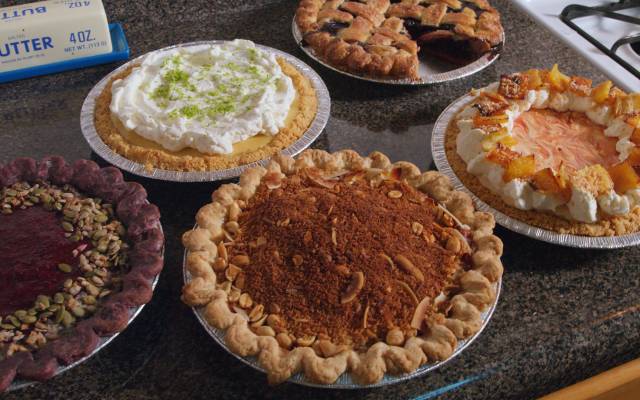Five assorted pies with various crusts and toppings on a kitchen countertop near butter and stovetop.