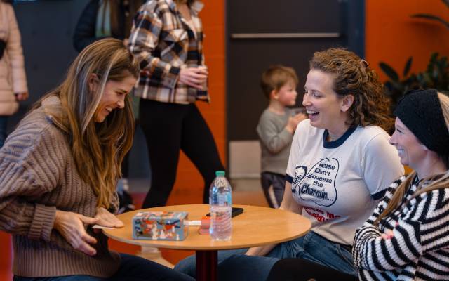 Three women sitting at a round table indoors with a water bottle, smartphone, and colorful box.