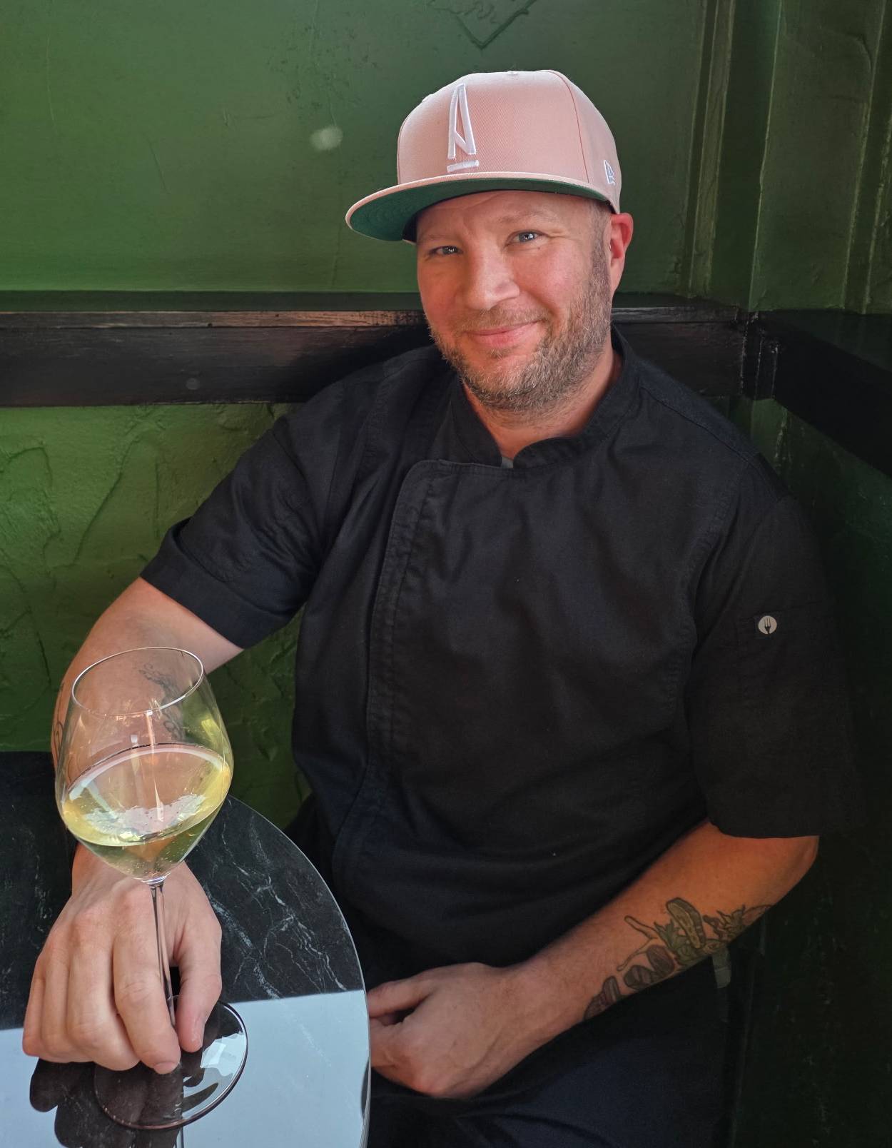 Man in black shirt and pink cap holding a glass of white wine at a black table near a green wall.