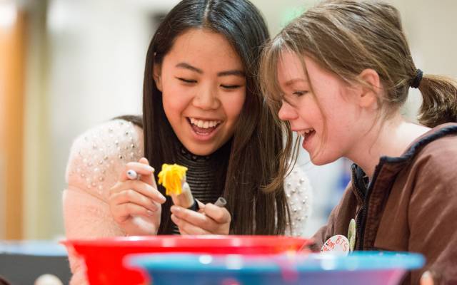 Two young women smiling and creating a craft using yellow yarn and a marker.