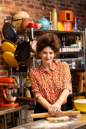 Woman rolling dough with a wooden rolling pin in a kitchen with brick wall and metal shelves.