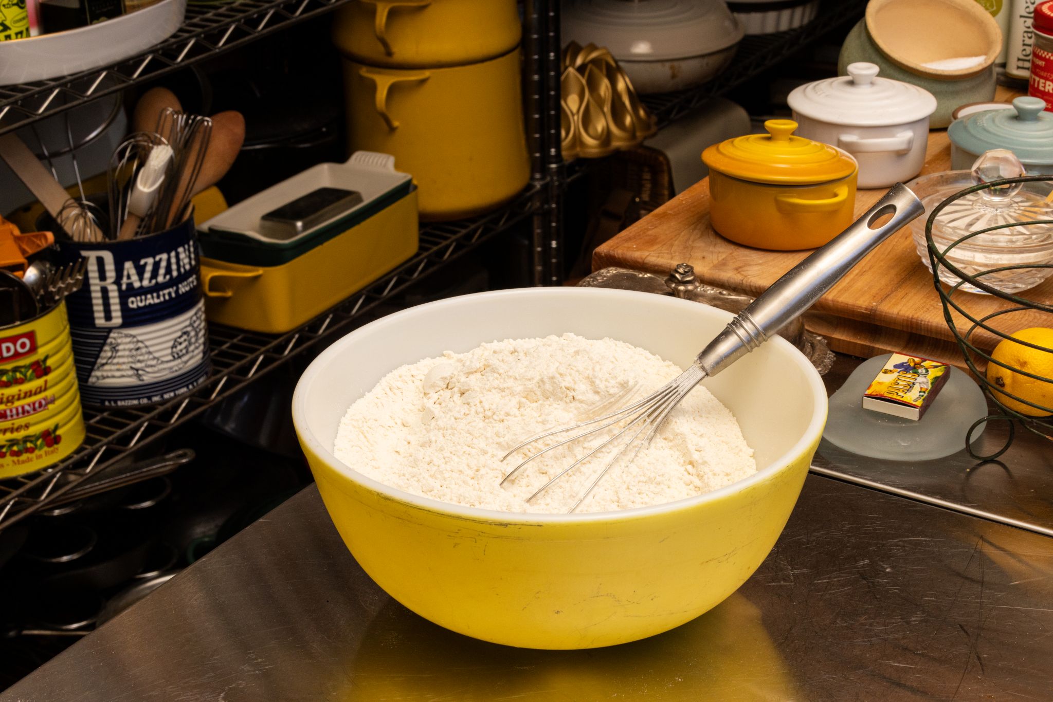 Yellow mixing bowl with flour and whisk on kitchen counter with utensils and containers.