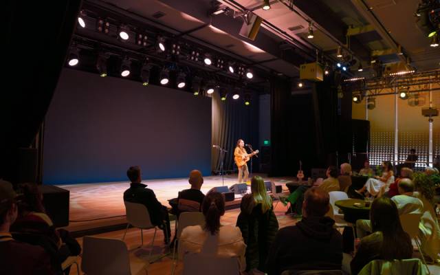 Musician playing acoustic guitar on stage with seated audience watching in a dimly lit venue