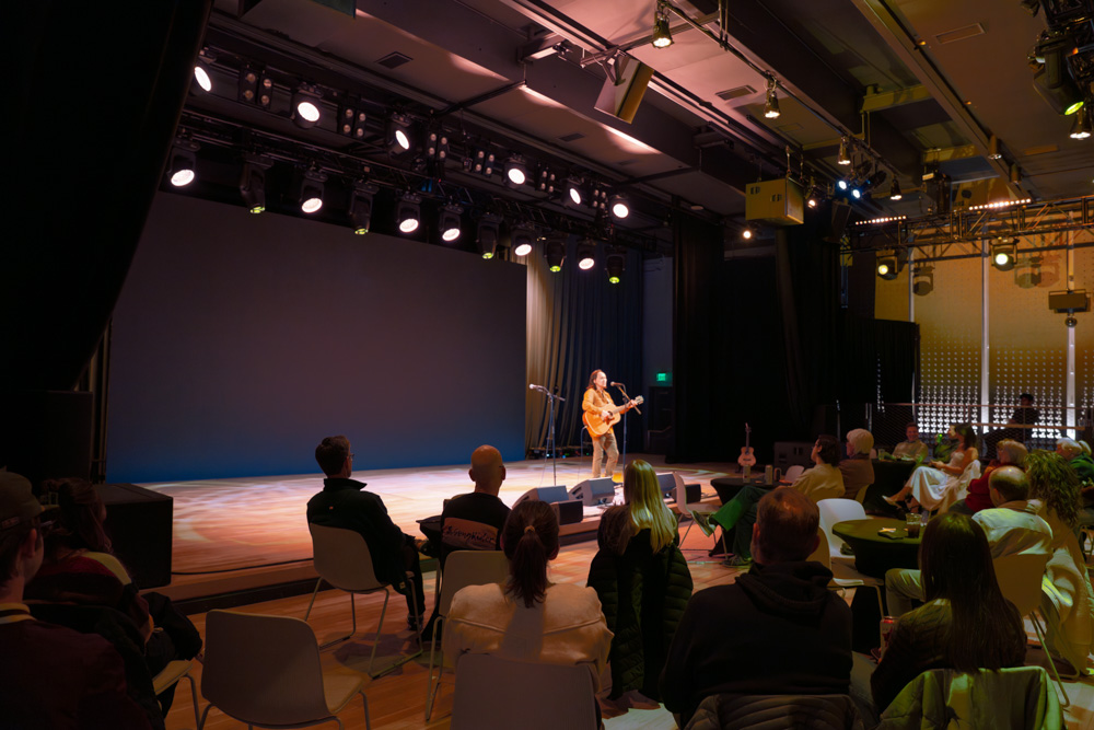 Musician playing acoustic guitar on stage with seated audience watching in a dimly lit venue