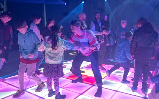 Adults and children dancing and interacting on an illuminated colorful indoor dance floor.