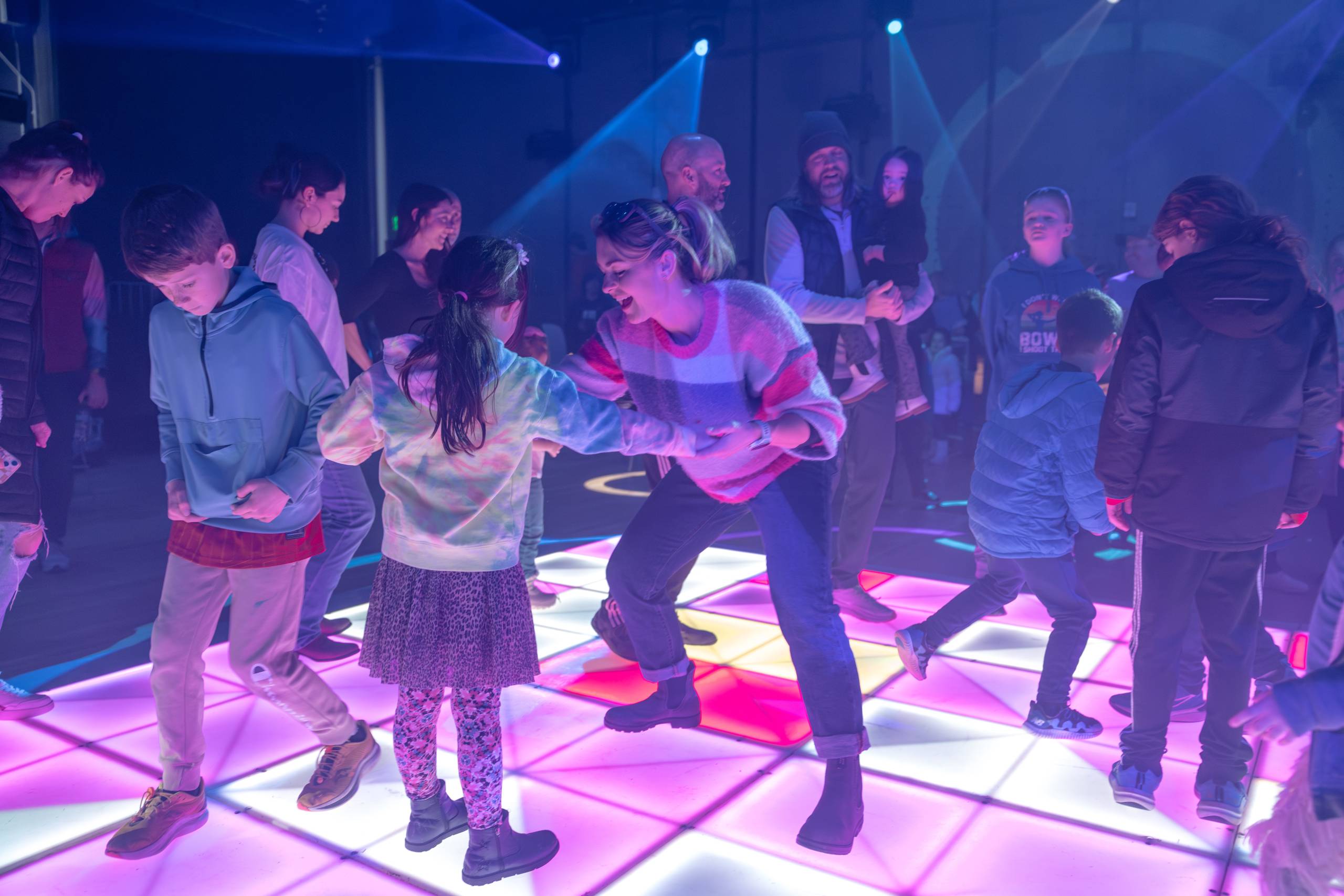 Adults and children dancing and interacting on an illuminated colorful indoor dance floor.