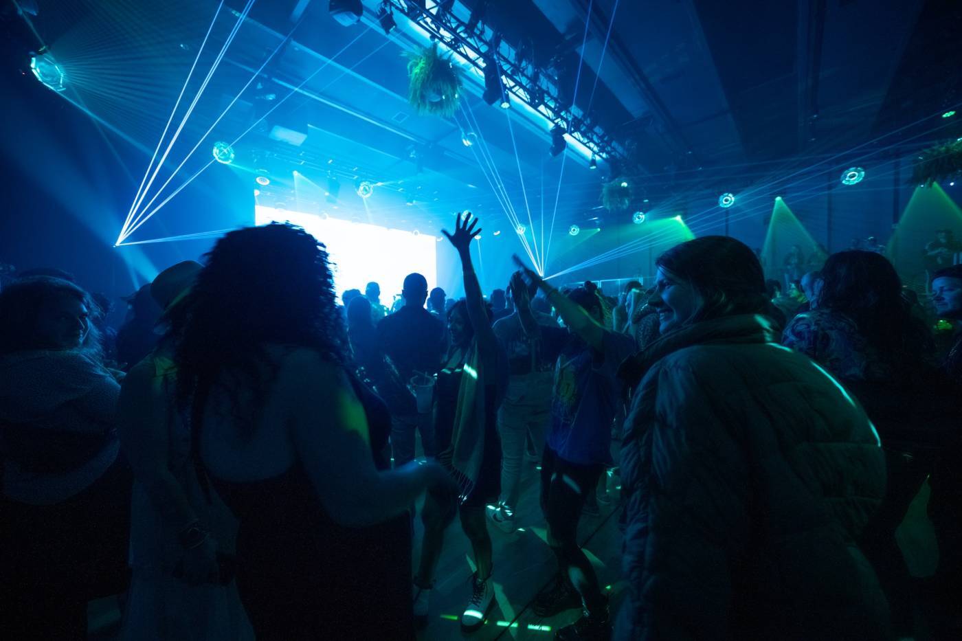 TGM08796-1400x933 Crowd dancing in a dimly lit venue with blue and green laser lights and raised hands.