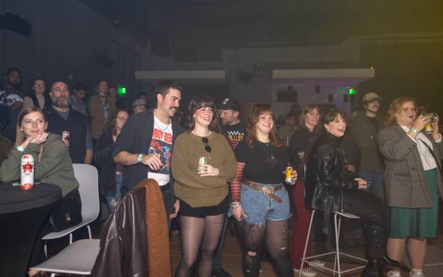Group of people indoors at a social event, some holding drinks and one taking a photo.