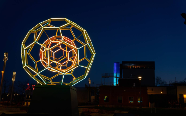 Geometric illuminated light sculpture with buildings and colorful vertical lights at night.