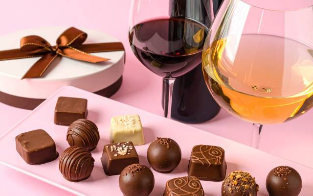 Box of assorted chocolates and two glasses of red and white wine on a pink surface