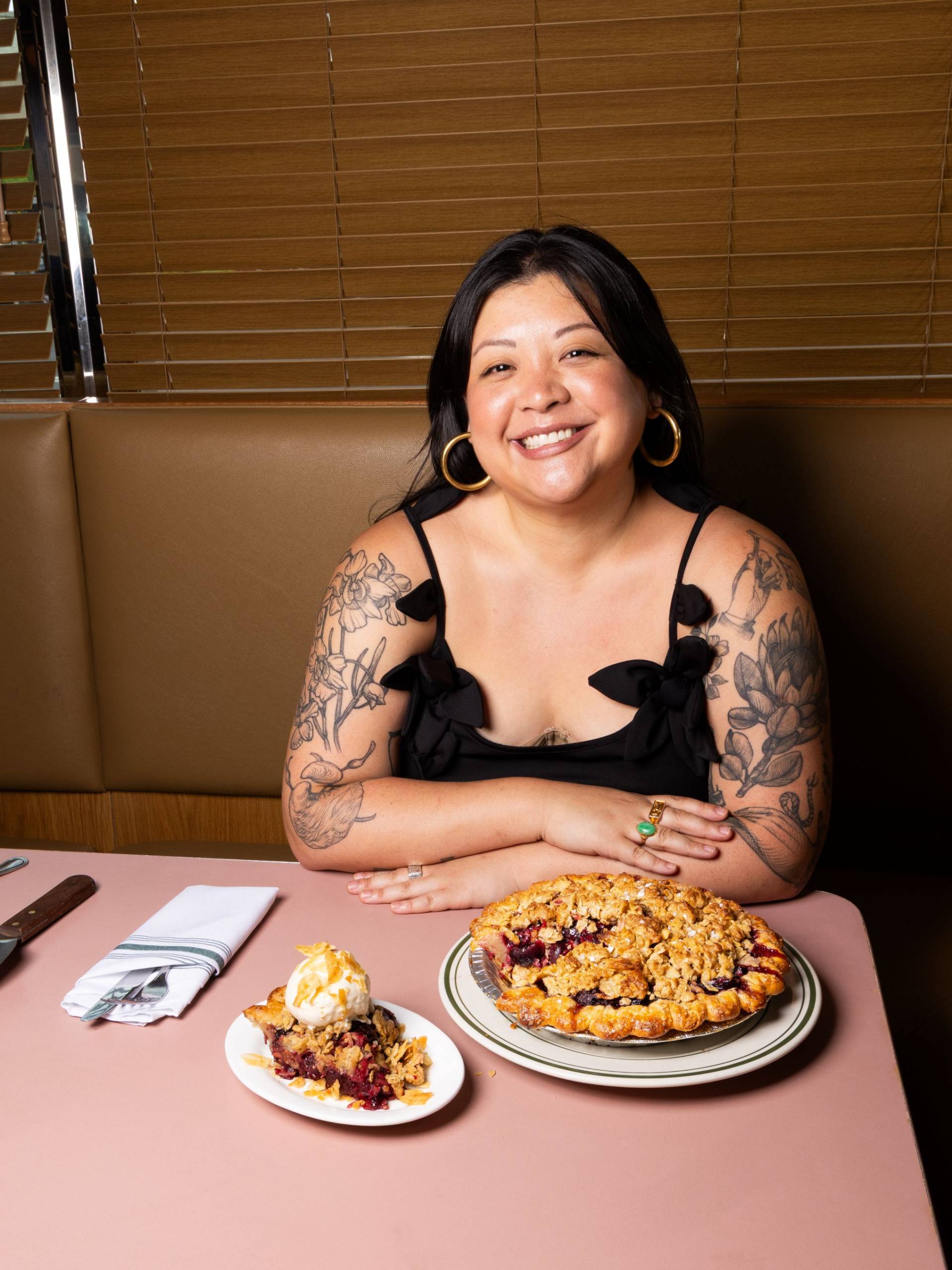 Woman with tattoos sitting at a table with a large fruit pie and a pie slice topped with ice cream.