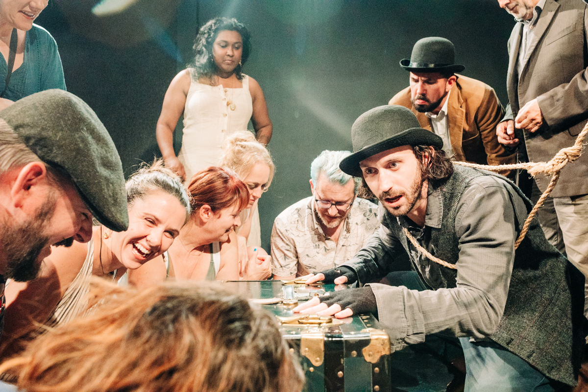 Group of people closely gathered around a large vintage trunk, with varied expressions and a rope involved.
