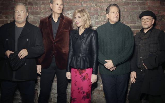 Five adults dressed in varied jackets and sweaters standing against a brick wall.