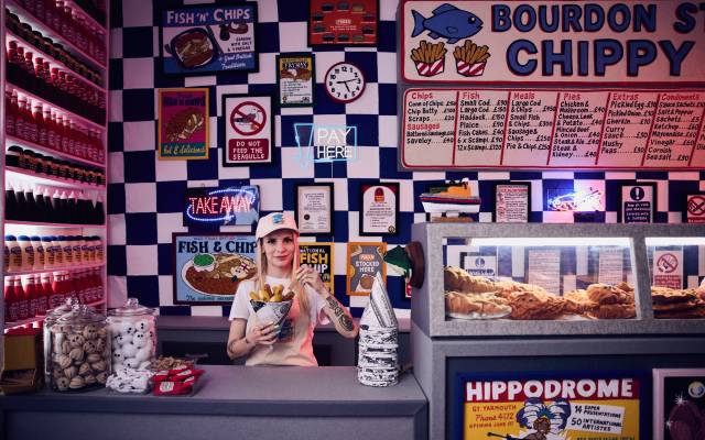 Counter of fish and chips shop with colorful signs, condiments, and a woman holding fries.