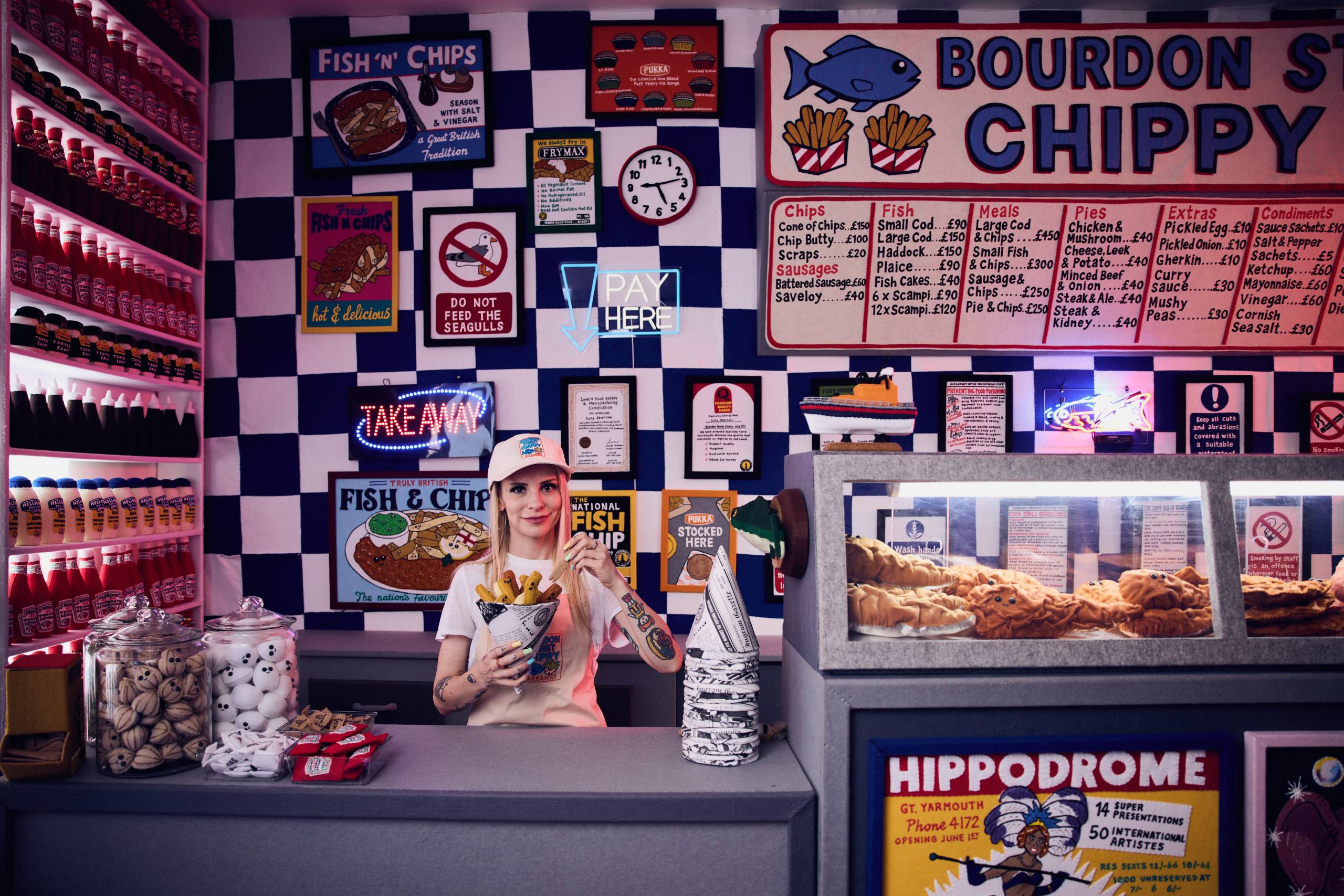 Counter of fish and chips shop with colorful signs, condiments, and a woman holding fries.
