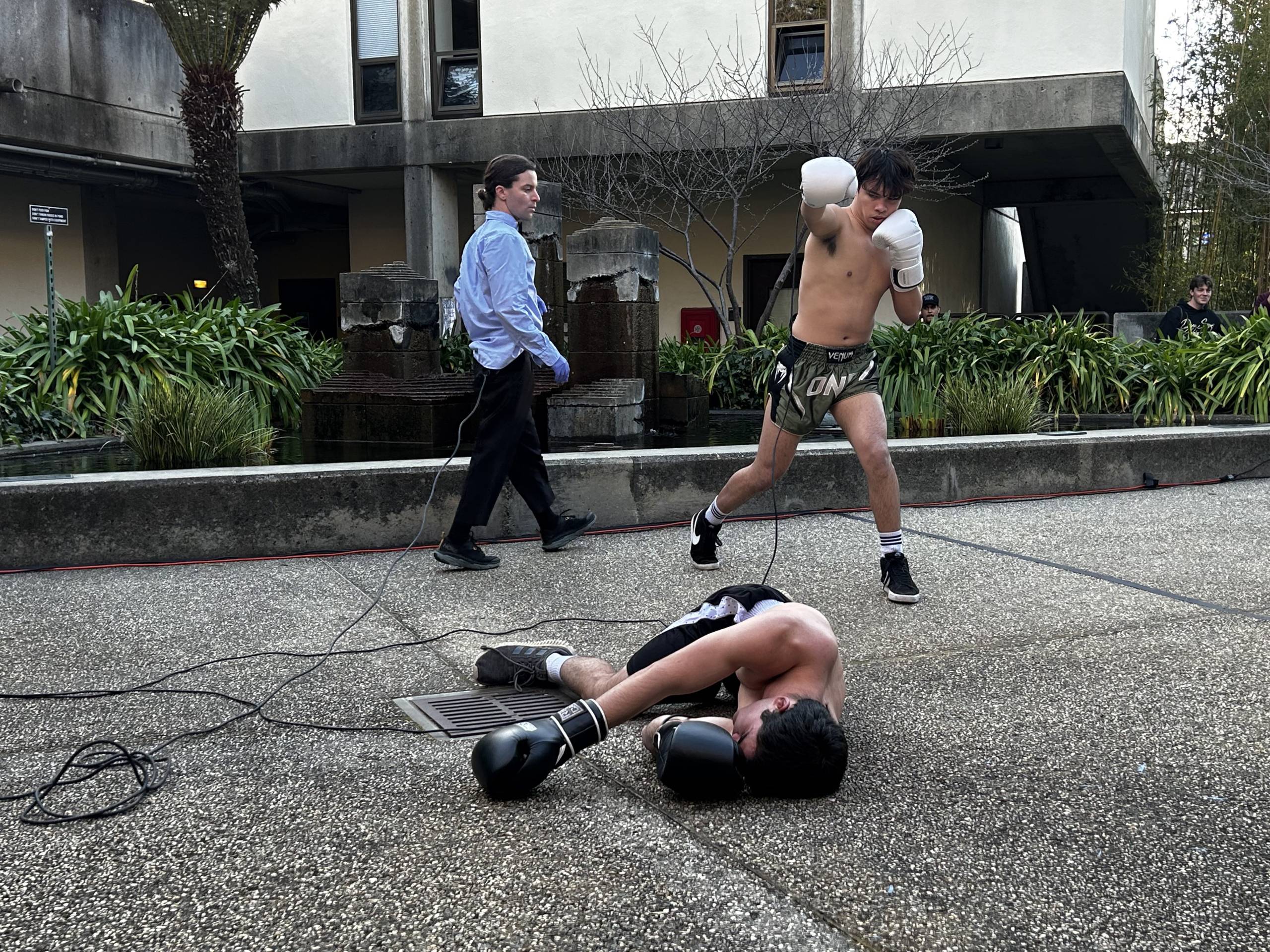 Two shirtless boxers fight on a paved outdoor area with a walking person nearby.