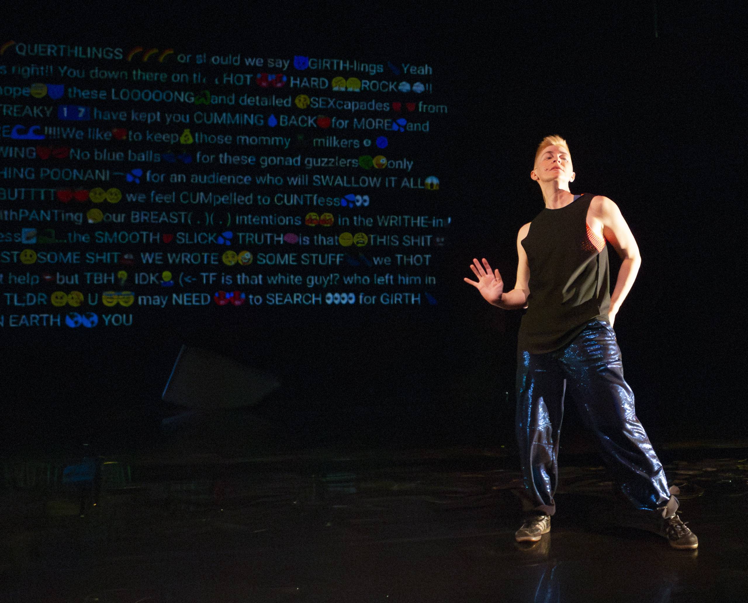Person in black sleeveless top and shiny blue pants stands on stage with colorful text projected behind.