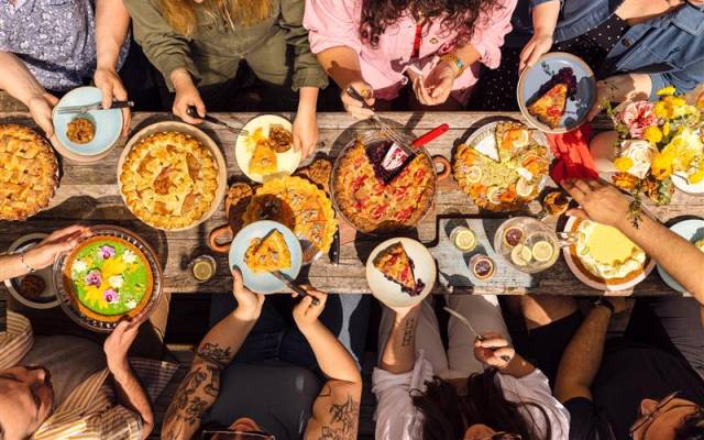 People sitting around a wooden table sharing various pies, desserts, drinks, and flowers.