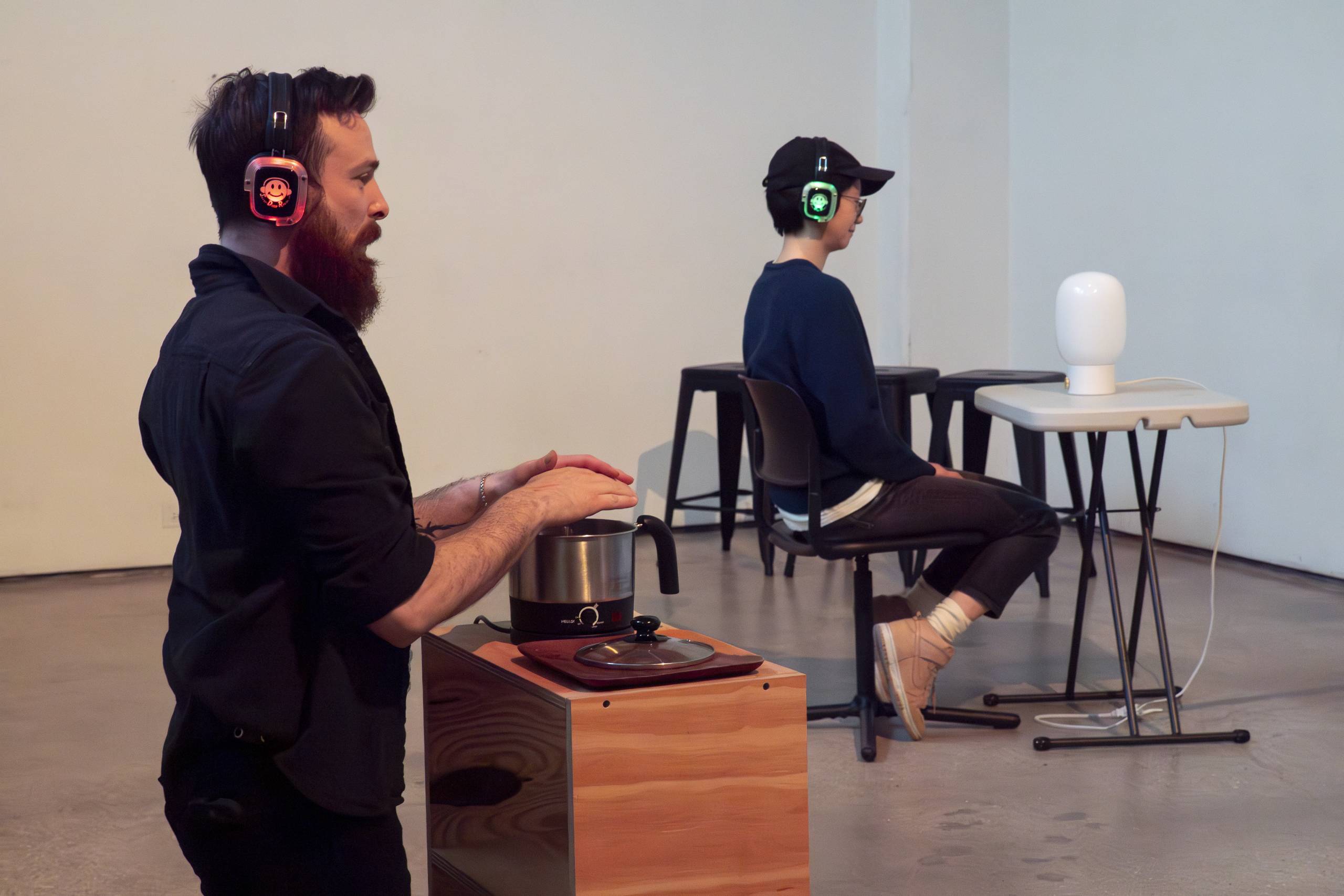 Two people wearing headphones interact with a heated pot and a white head-shaped object indoors.
