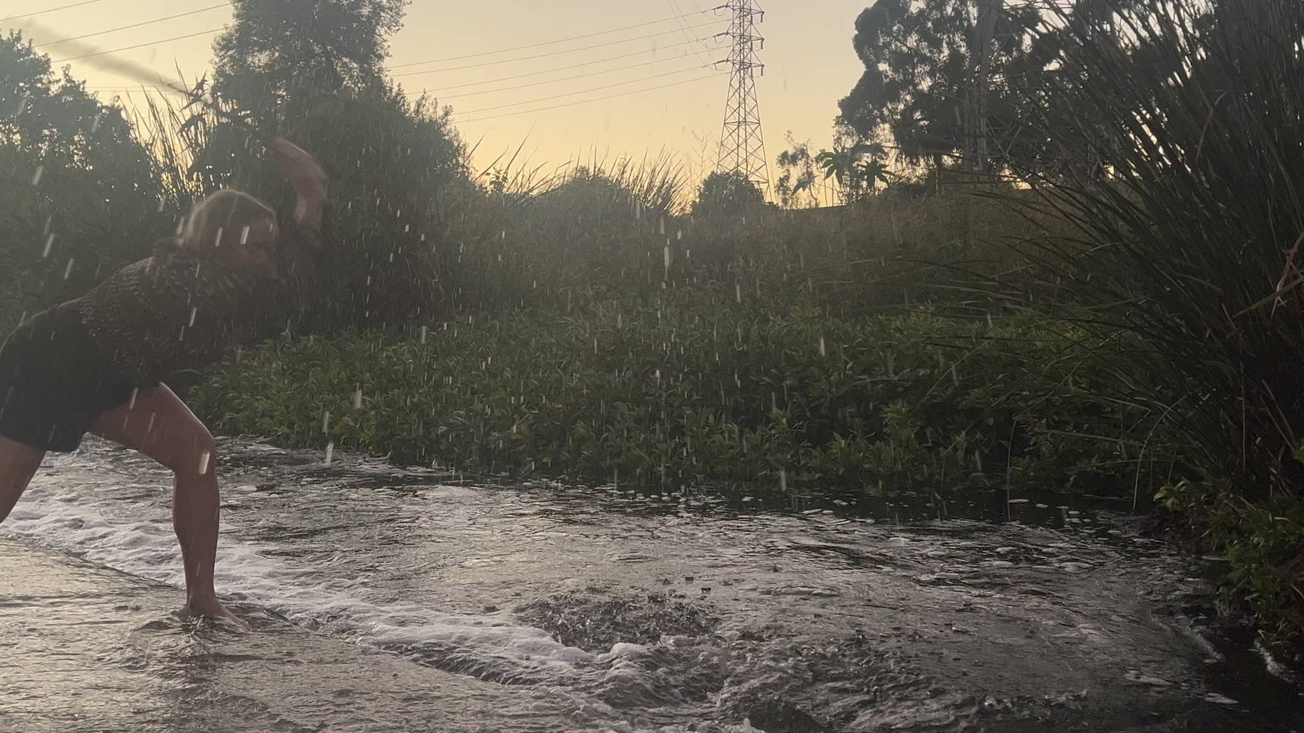 Person splashing water in a shallow stream with vegetation and power lines in the background at dusk