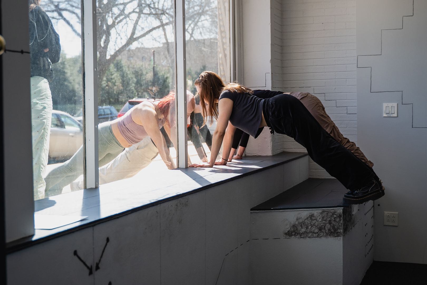 Two people doing push-ups indoors by a sunlit large window with a third person standing nearby