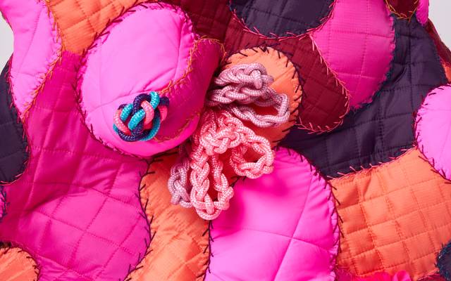 Quilted fabric artwork with pink, orange, and dark red patches featuring knotted and braided cords.