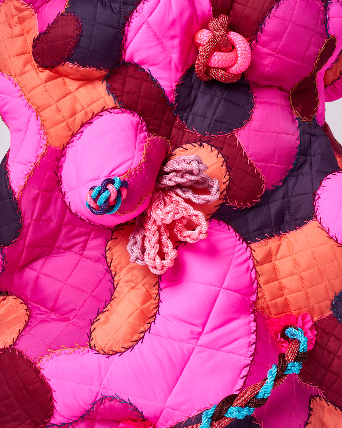 Quilted fabric artwork with pink, orange, and dark red patches featuring knotted and braided cords.