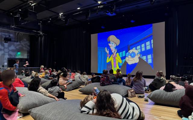 Children and adults seated on gray cushions watching a cartoon on a large screen in a dark room.
